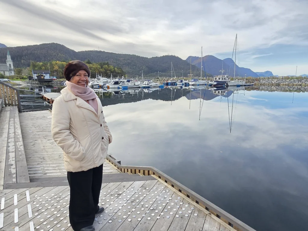 Danny's mother smiling on a wooden pier next to a peaceful harbor reflecting boats, mountains, and a village church in Ålesund during a Holland America Rotterdam shore excursion. Things to do in Ålesund from cruise ship, by Danny and Peter.