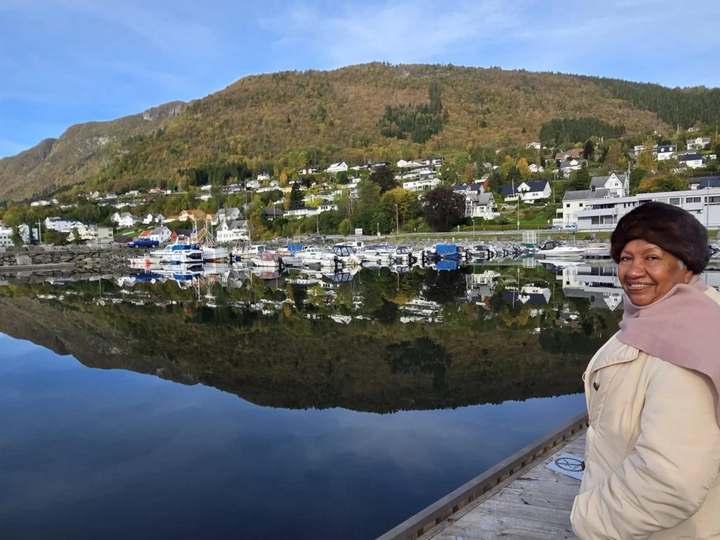 Danny's mother smiling on a pier next to a serene harbor reflecting hillside village homes, autumn trees, and docked boats near Ålesund during a Holland America Rotterdam shore excursion. Things to do in Ålesund from cruise ship, by Danny and Peter.