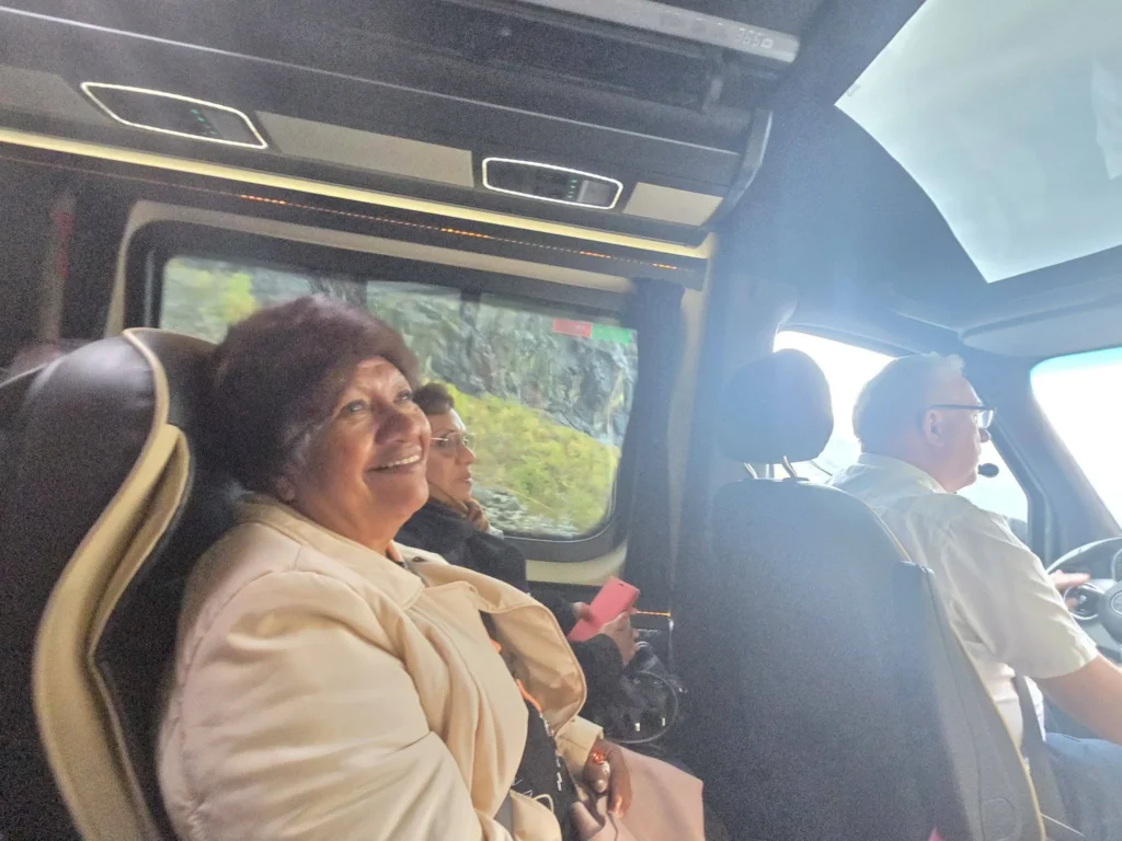 Danny's mother smiling brightly while seated next to Danny's aunt inside a tour bus as the driver navigates through scenic mountain roads near Ålesund during a Holland America Rotterdam shore excursion. Things to do in Ålesund from cruise ship, by Danny and Peter.