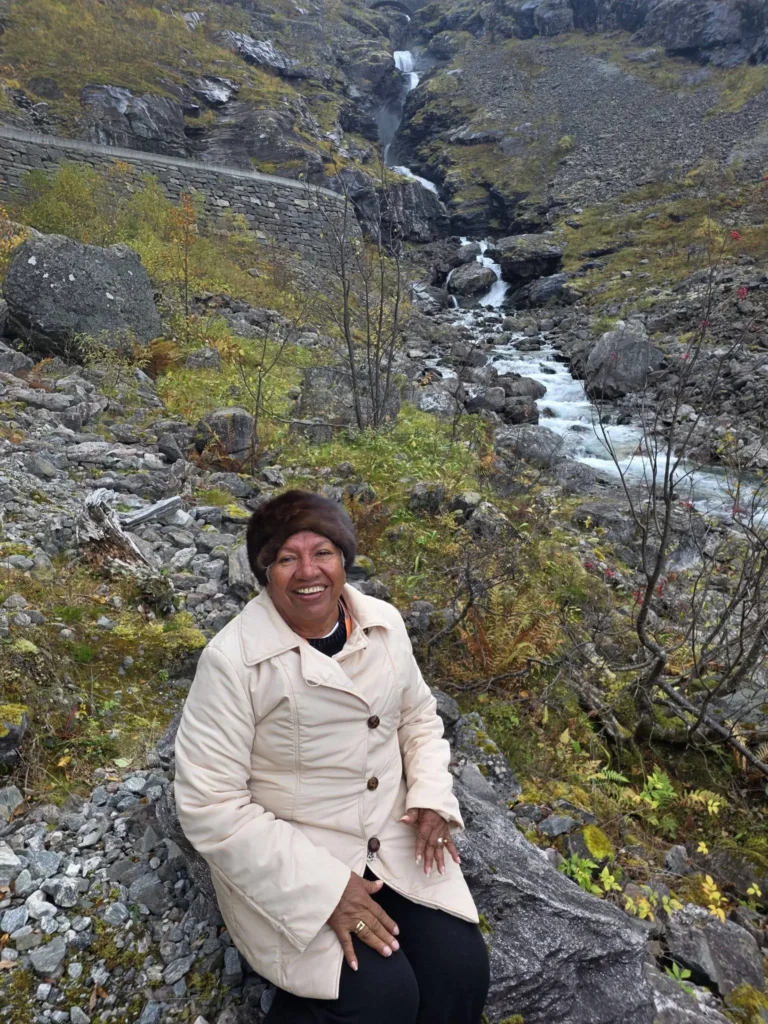 Danny's mother smiling warmly while sitting on a boulder in front of a cascading mountain stream and rocky waterfall near Ålesund during a Holland America Rotterdam shore excursion. Things to do in Ålesund from cruise ship, by Danny and Peter.