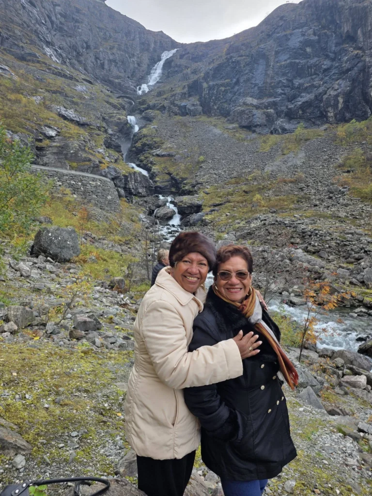 Danny's mother hugging Danny's aunt while smiling together in front of Stigfossen waterfall cascading down dramatic mountain cliffs near Ålesund during a Holland America Rotterdam shore excursion. Things to do in Ålesund from cruise ship, by Danny and Peter.
