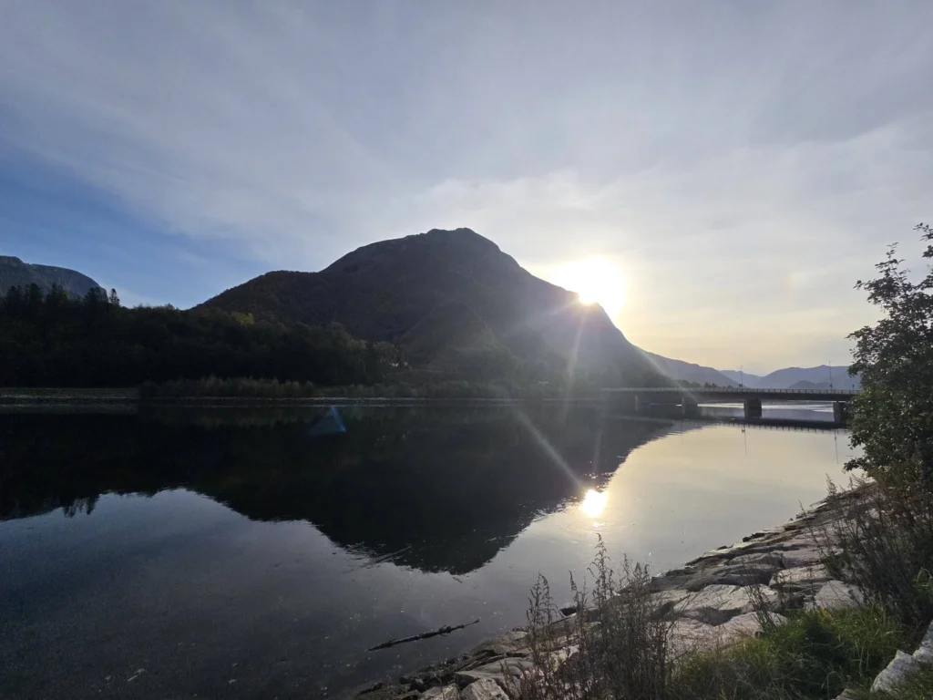 The sun peeking over a silhouetted mountain peak, casting bright rays and reflections across calm waters and a distant bridge near Ålesund during a Holland America Rotterdam shore excursion. Things to do in Ålesund from cruise ship, by Danny and Peter.