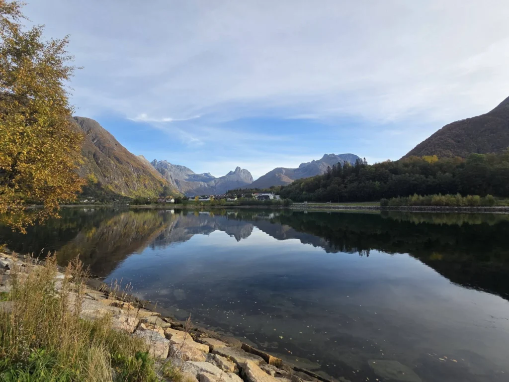 Calm waters reflecting dramatic alpine mountains, autumn foliage, and blue skies framed by golden trees along a scenic drive near Ålesund during a Holland America Rotterdam shore excursion. Things to do in Ålesund from cruise ship, by Danny and Peter.