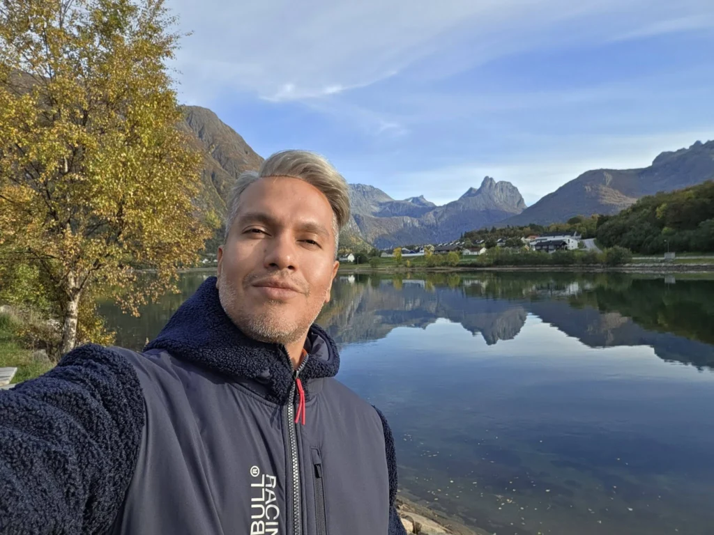 Danny taking a selfie in front of a calm fjord lake reflecting dramatic mountain peaks and autumn trees near Ålesund during a Holland America Rotterdam shore excursion. Things to do in Ålesund from cruise ship, by Danny and Peter.
