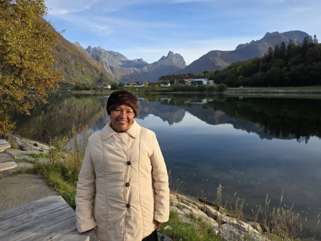 Danny's mother smiling warmly beside a calm fjord lake reflecting sharp mountain peaks and autumn foliage near Ålesund during a Holland America Rotterdam shore excursion. Things to do in Ålesund from cruise ship, by Danny and Peter.