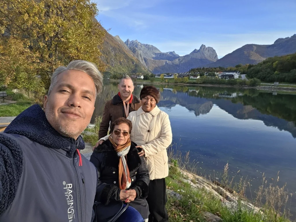 Group selfie taken alongside a placid lake during a scenic valley drive in Ålesund, Norway, with jagged mountain reflections mirroring in the clear water under a sunny autumn sky. Things to do in Ålesund from cruise ship, by Danny and Peter.