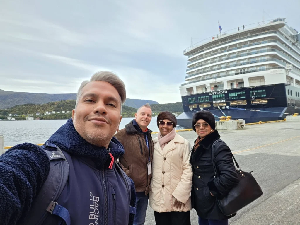 Group selfie taken on Storneskaia Quay in front of the stern of the Holland America Line cruise ship Rotterdam docked in Ålesund, Norway, with surrounding mountains in the background. Rotterdam Ålesund Docking Location, by Danny and Peter.