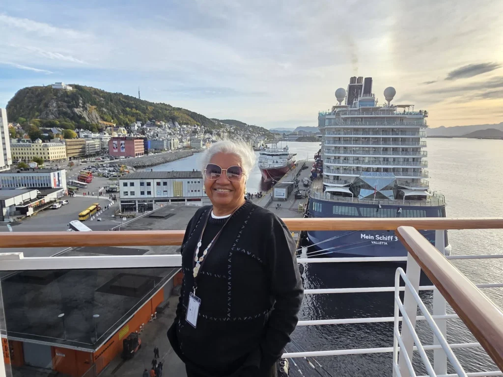 Danny's mother smiling on an upper cruise deck looking over Storneskaia Quay in Ålesund, Norway, with the docked Mein Schiff 3 and Mount Aksla in the background. Ålesund Norway Cruise Port Guide, by Danny and Peter.