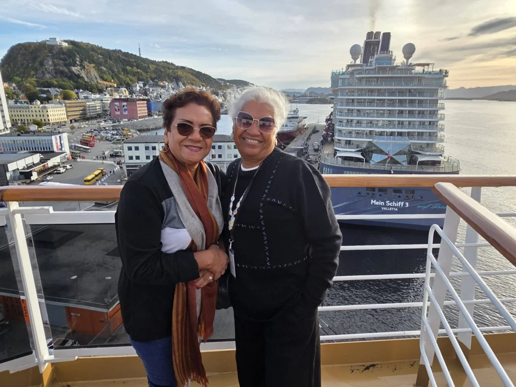 Danny's mother and aunt smiling together on an upper cruise ship railing at Storneskaia Quay in Ålesund, Norway, with cruise ship Mein Schiff 3 docked alongside and Mount Aksla in the background. Ålesund Norway Cruise Port Guide, by Danny and Peter.