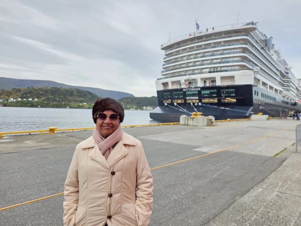 Danny's mother standing on Storneskaia Quay in front of Holland America Line's Rotterdam docked in Ålesund, Norway, with forested mountains across the water in the background. MS Rotterdam Ålesund Docking Location, by Danny and Peter.