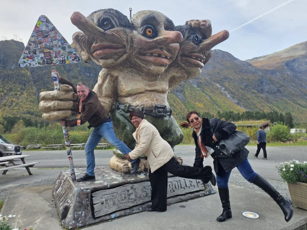 Peter along with Danny's mother and aunt playfully posing in front of a giant three-headed troll statue with a road sign at Trollstigen Gjestegård near Ålesund, Norway. Things to do in Ålesund from cruise ship, by Danny and Peter.