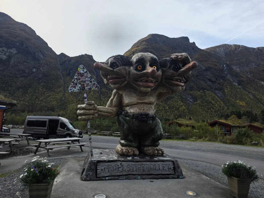 A large three-headed troll statue holding a stickered road sign at Trollstigen Gjestegård, set against dramatic steep mountains near Ålesund, Norway. Things to do in Ålesund from cruise ship, by Danny and Peter.
