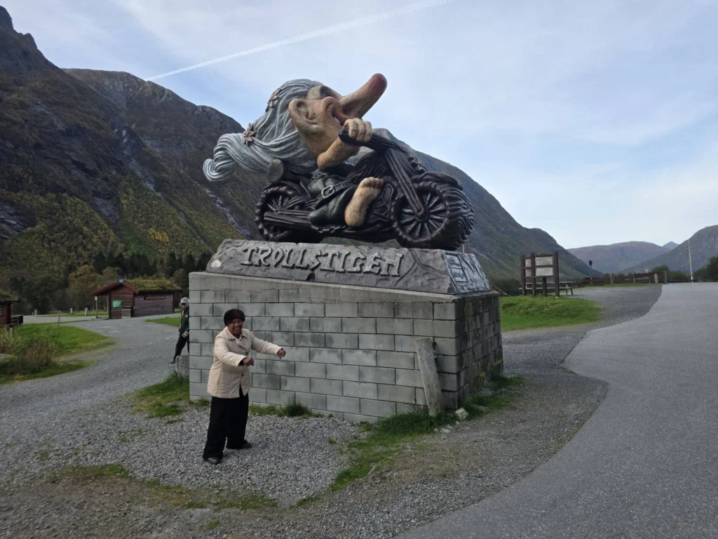 Danny's mother playfully posing below a large statue of a troll riding a motorcycle at Trollstigen Gjestegård near Ålesund, Norway, with steep mountains in the background. Things to do in Ålesund from cruise ship, by Danny and Peter.