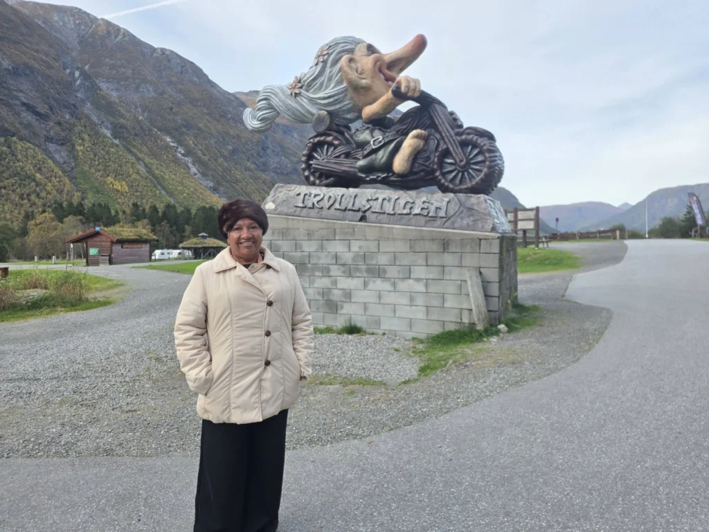 Danny's mother smiling standing in front of the motorcycle troll statue at Trollstigen Gjestegård near Ålesund, Norway, with steep green mountains in the background. Things to do in Ålesund from cruise ship, by Danny and Peter.