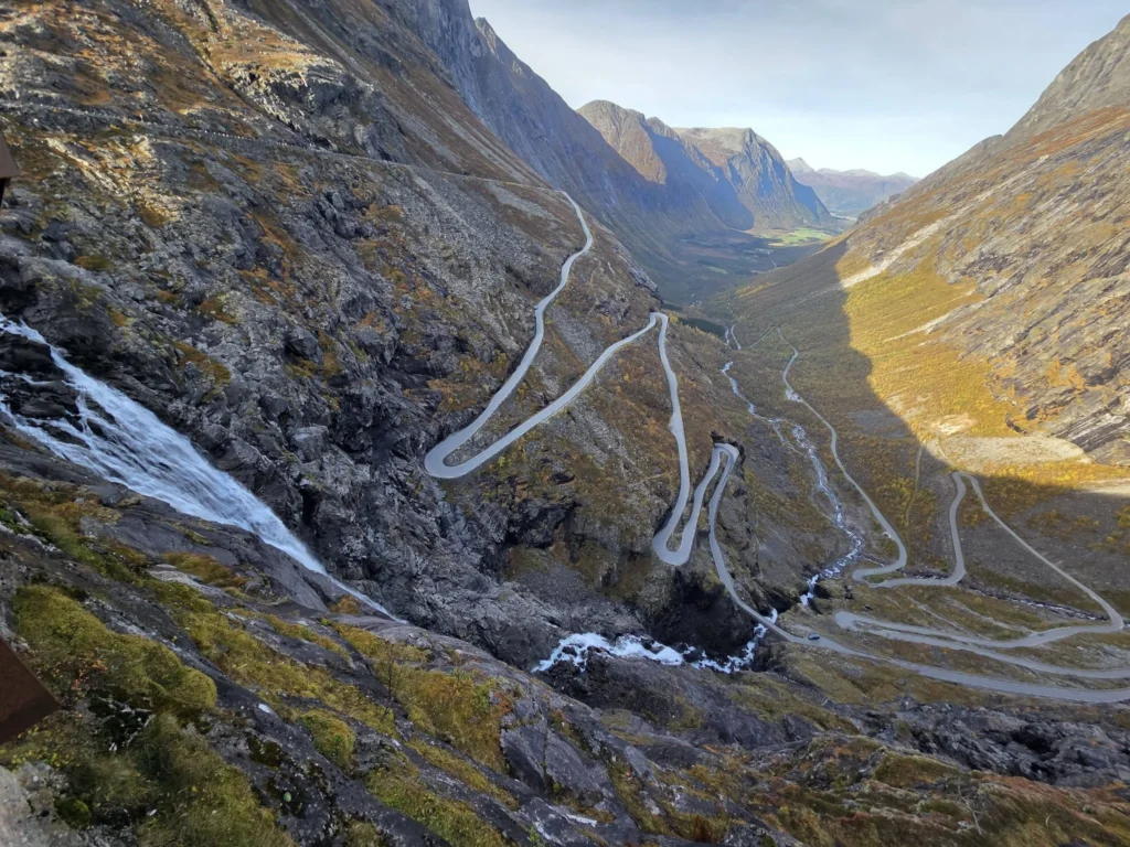 Panoramic high-angle view of the iconic winding hairpin bends of the Trollstigen mountain road carving through a deep valley with Stigfossen waterfall cascading down rocky cliffs near Ålesund, Norway. Things to do in Ålesund from cruise ship, by Danny and Peter.