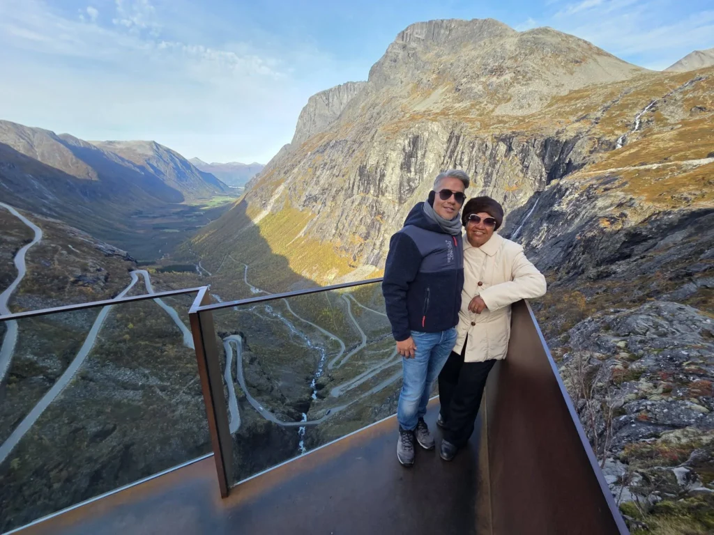Danny and his mother standing on the glass viewing platform at Trollstigen with towering mountain cliffs and the winding road below near Ålesund, Norway. Things to do in Ålesund from cruise ship, by Danny and Peter.