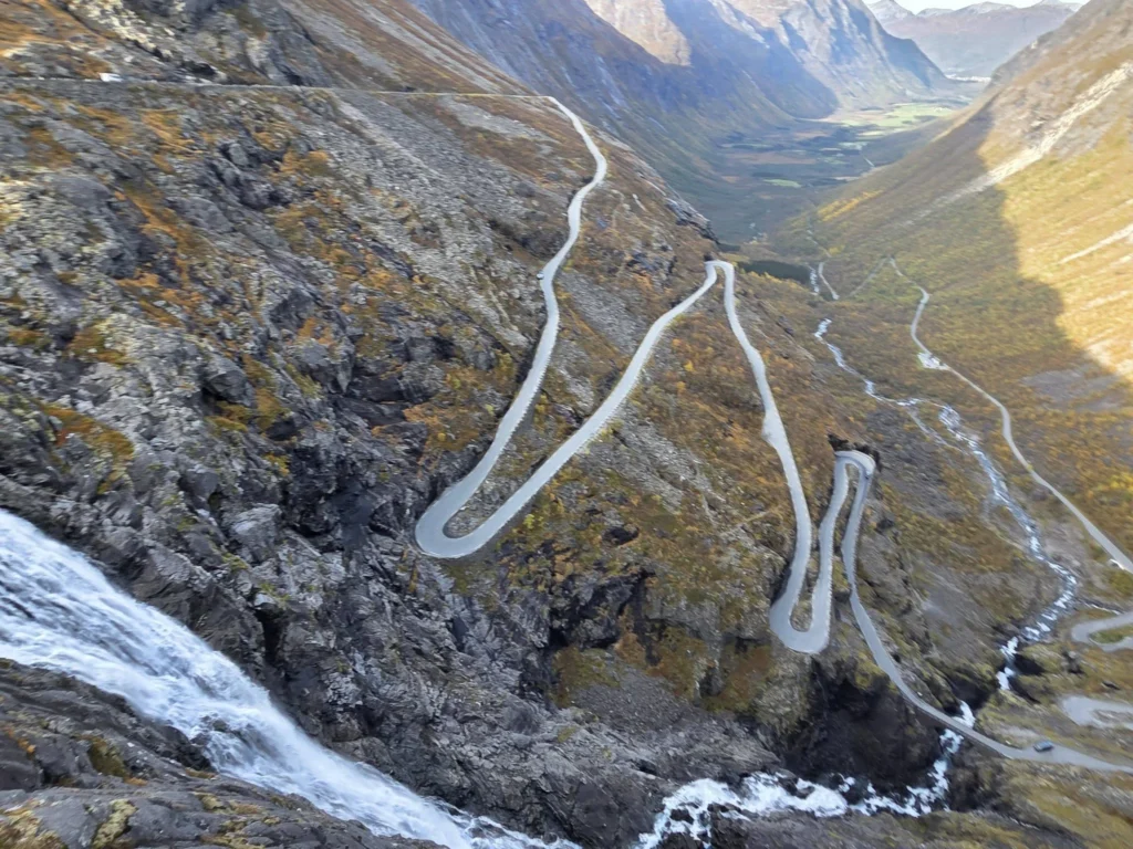 High-angle perspective showing Stigfossen waterfall rushing past rocky cliffs alongside the steep, winding hairpin turns of Trollstigen near Ålesund, Norway. Things to do in Ålesund from cruise ship, by Danny and Peter.