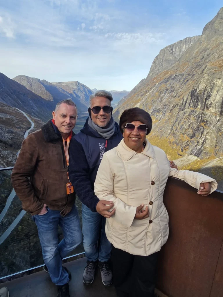 Peter, Danny, and Danny's mother standing together on the Trollstigen viewing platform with steep mountain cliffs and the winding pass in the background near Ålesund, Norway. Things to do in Ålesund from cruise ship, by Danny and Peter.