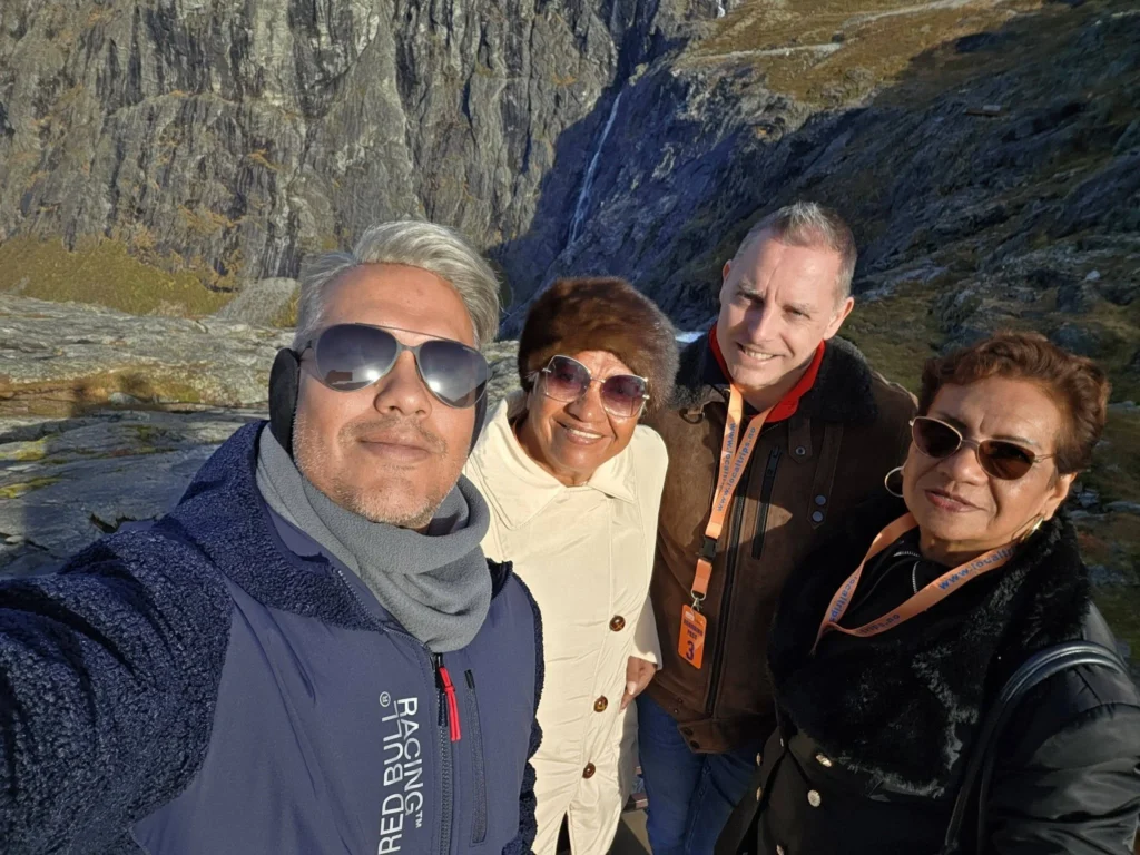 Group selfie of Danny, Danny's mother, Peter, and Danny's aunt smiling together at the Trollstigen viewpoint plateau with rocky mountain cliffs in the background near Ålesund, Norway. Things to do in Ålesund from cruise ship, by Danny and Peter.