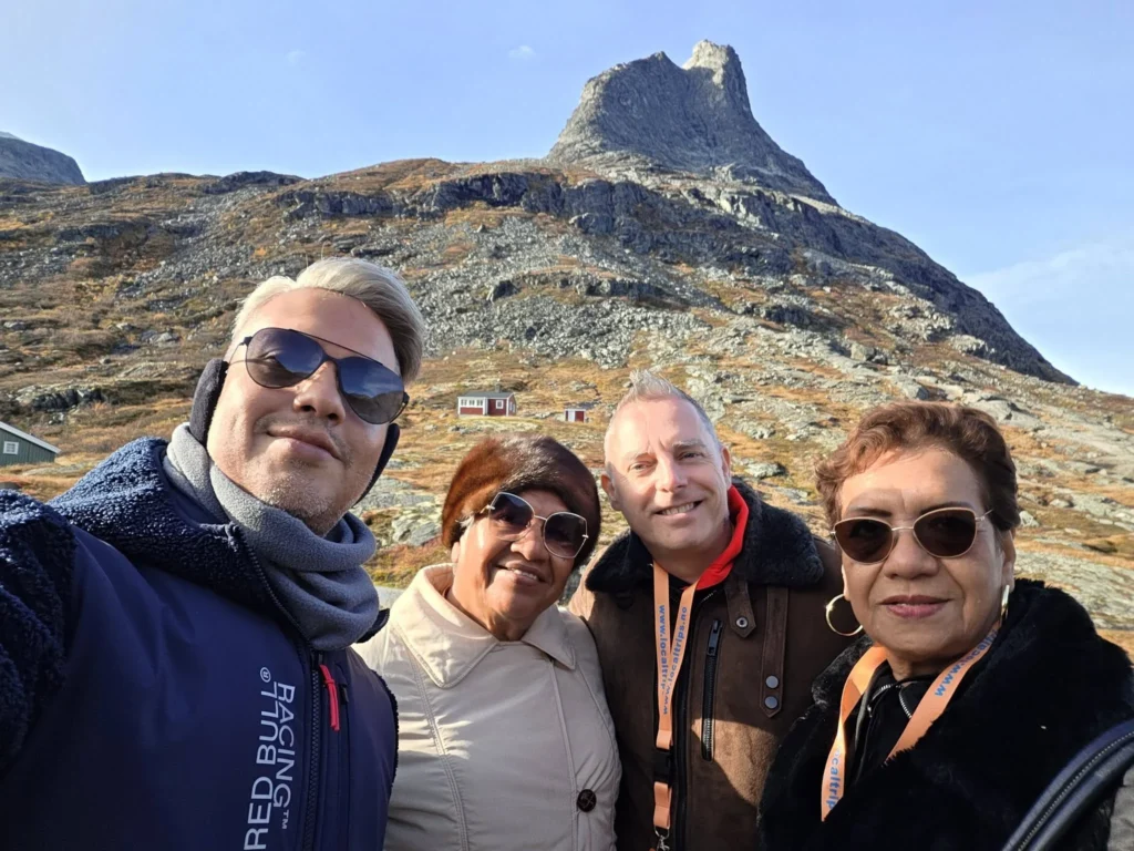 Group selfie of Danny, Danny's mother, Peter, and Danny's aunt outdoors at the Trollstigen plateau with a sharp rocky mountain peak reaching into the clear blue sky near Ålesund, Norway. Things to do in Ålesund from cruise ship, by Danny and Peter.