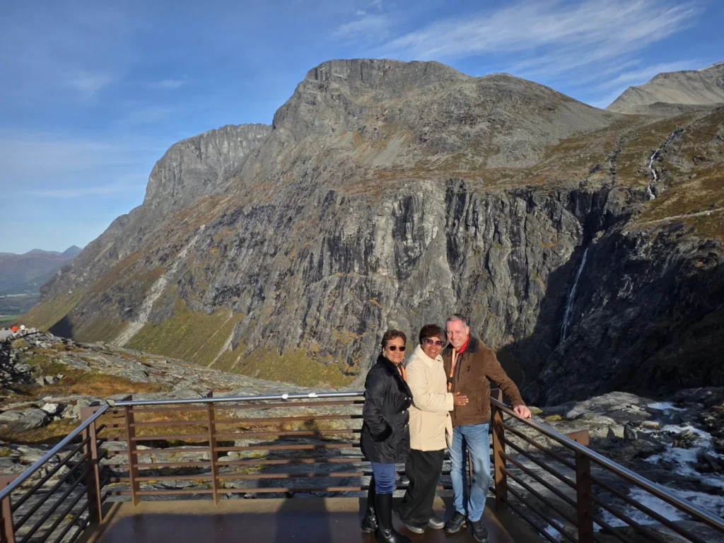 Danny's aunt, Danny's mother, and Peter standing on an outdoor observation deck at the Trollstigen plateau with massive rocky mountains and a distant waterfall near Ålesund, Norway. Things to do in Ålesund from cruise ship, by Danny and Peter.