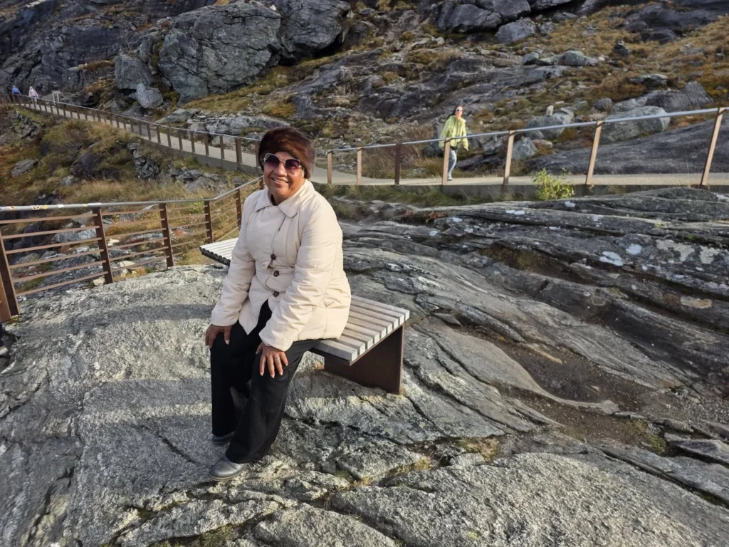 Danny's mother smiling while resting on a wooden bench along the rocky walking paths at the Trollstigen viewpoint plateau near Ålesund, Norway. Things to do in Ålesund from cruise ship, by Danny and Peter.