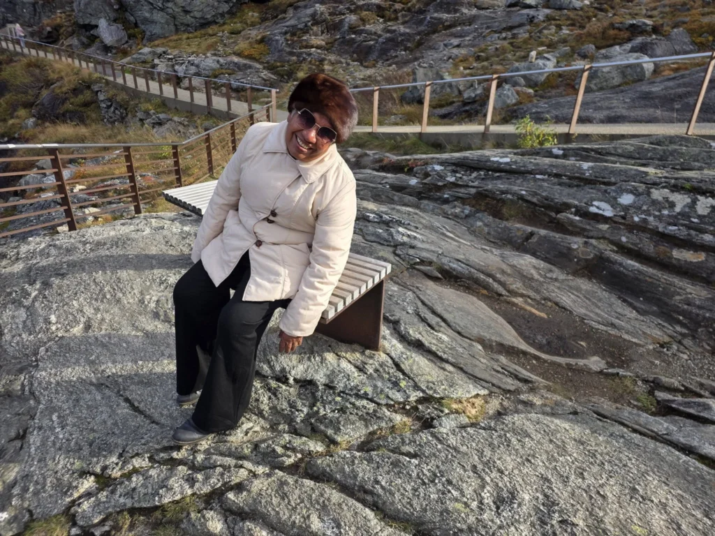 Danny's mother happily leaning back while sitting on a wooden bench along the rocky paths of the Trollstigen viewpoint plateau near Ålesund, Norway. Things to do in Ålesund from cruise ship, by Danny and Peter.