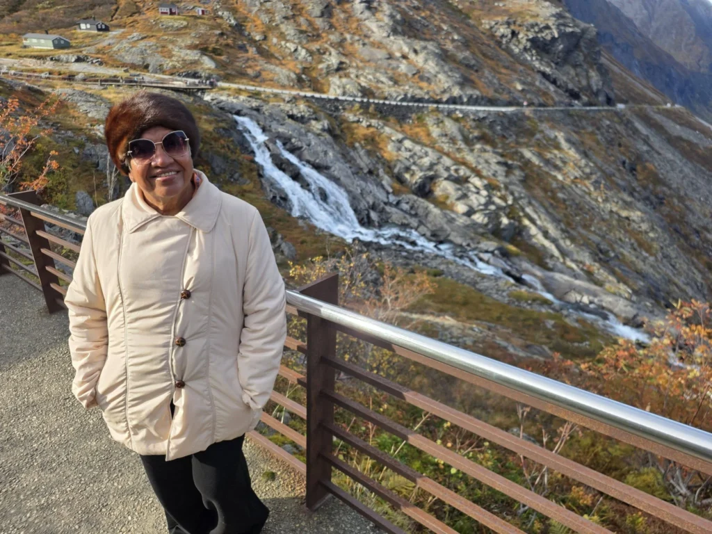Danny's mother smiling along a railed walkway at the Trollstigen viewpoint plateau with a mountain stream flowing down rocky slopes near Ålesund, Norway. Things to do in Ålesund from cruise ship, by Danny and Peter.