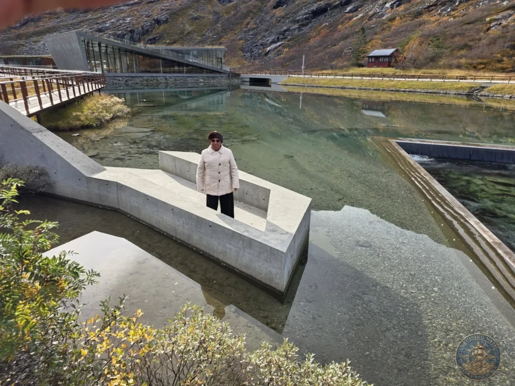 Danny's mother standing on a sleek concrete walkway extension over the clear reflecting pool at the modern Trollstigen visitor center plateau near Ålesund, Norway. Things to do in Ålesund from cruise ship, by Danny and Peter.